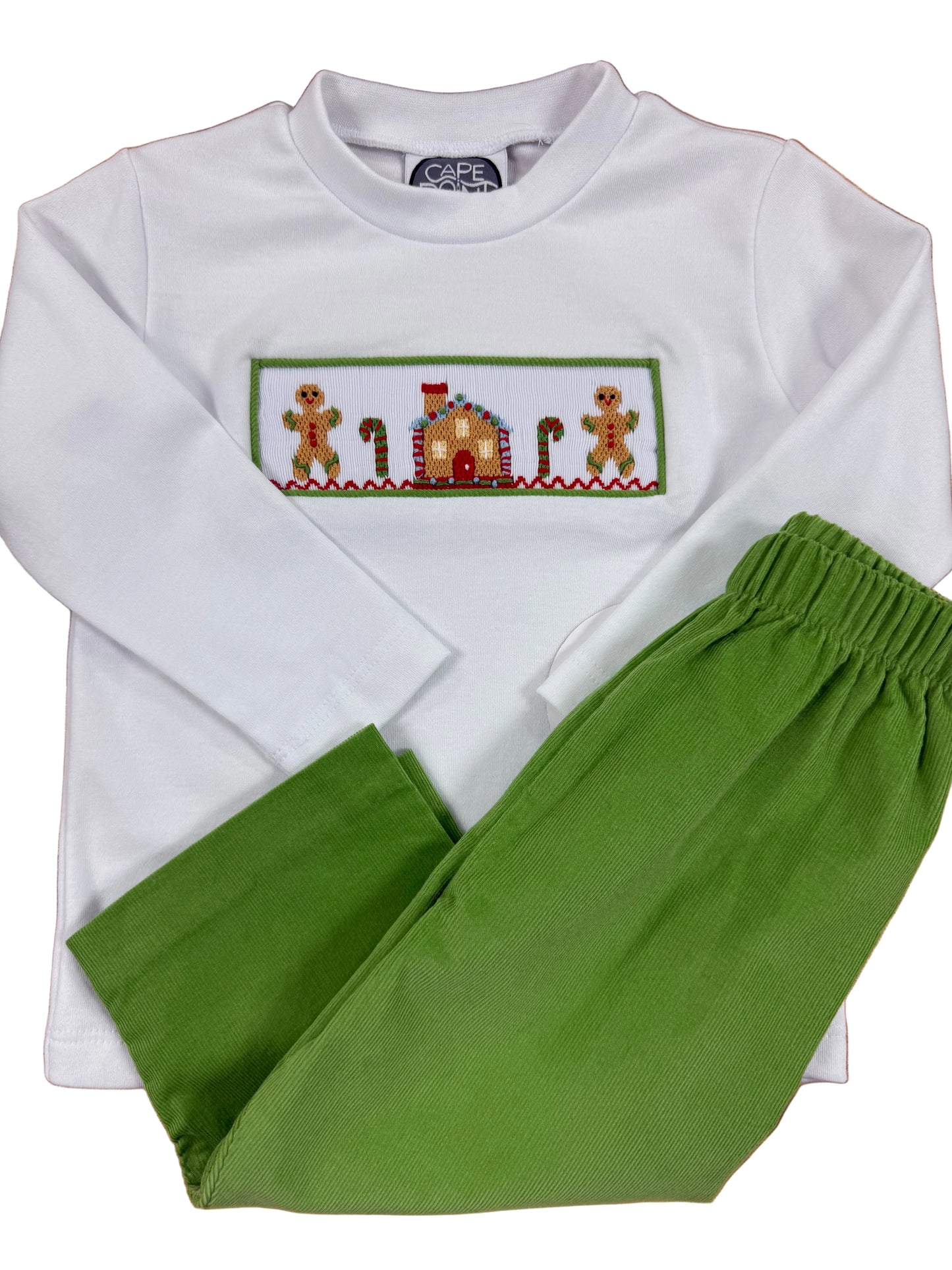 A children's holiday-themed two-piece pajama set laid flat on a wooden floor, featuring a white long-sleeve top with a colorful embroidered gingerbread house and gingerbread men across the chest, paired with green pants.