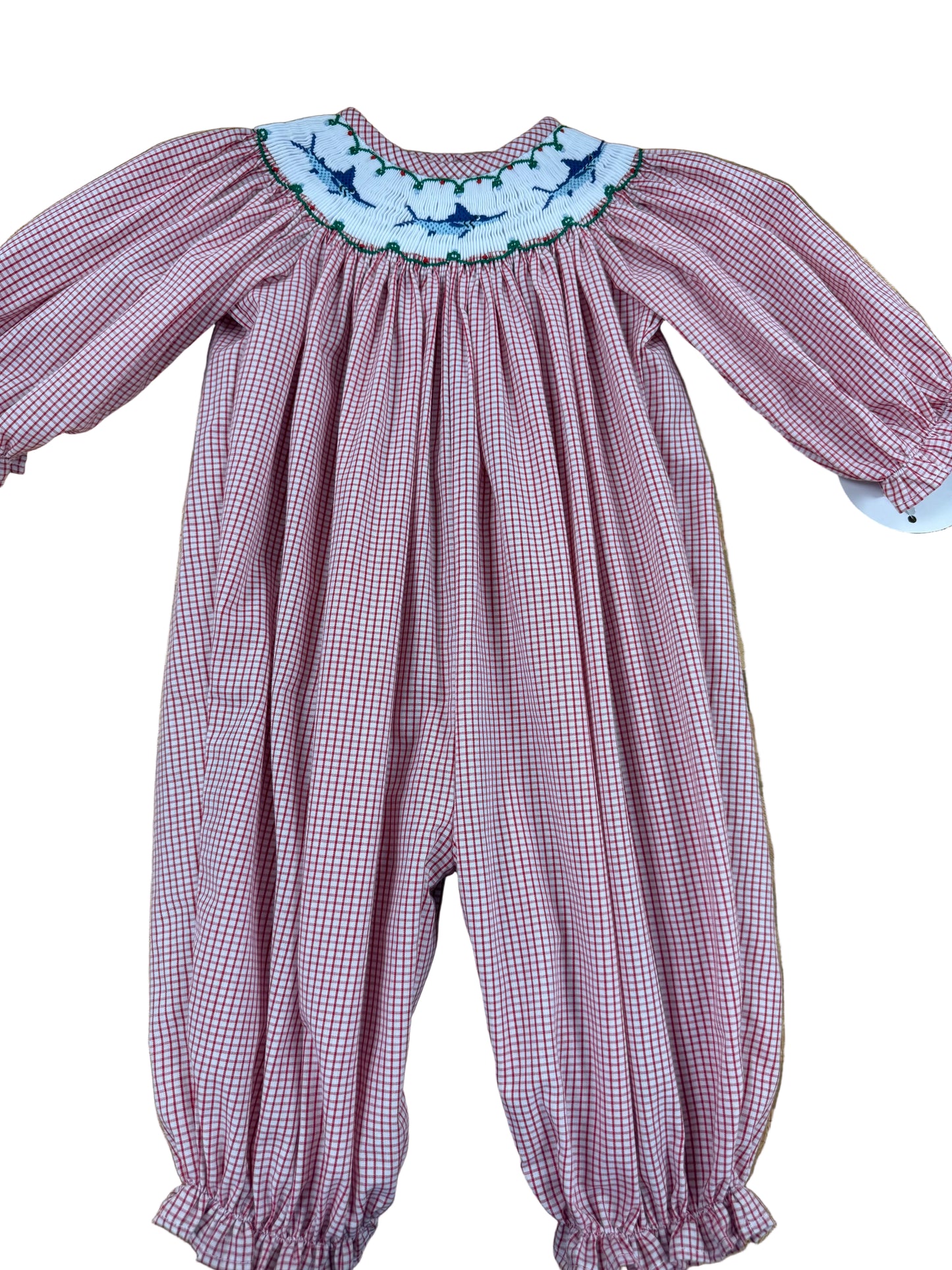 A baby smocked romper in a pink and white gingham pattern, featuring a white floral embroidered collar with blue and green accents, laid flat on a wooden floor.