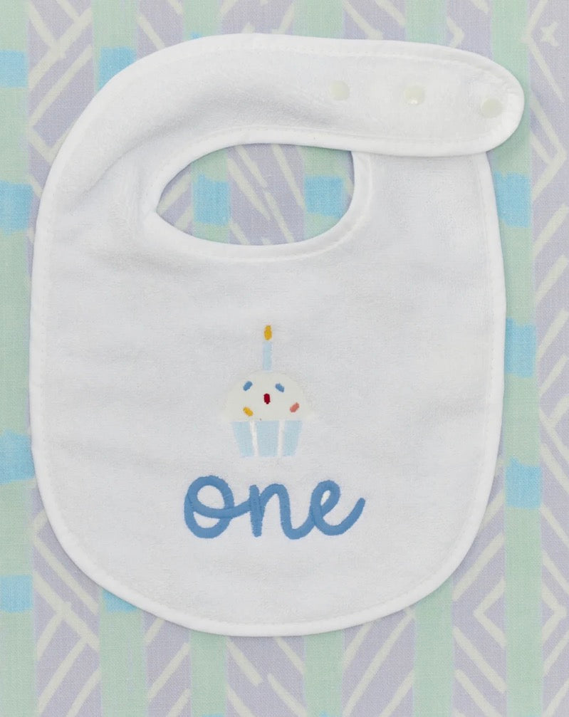 A white baby bib with a cupcake graphic and the word 'one' in blue script, laid flat on a pastel patterned background.