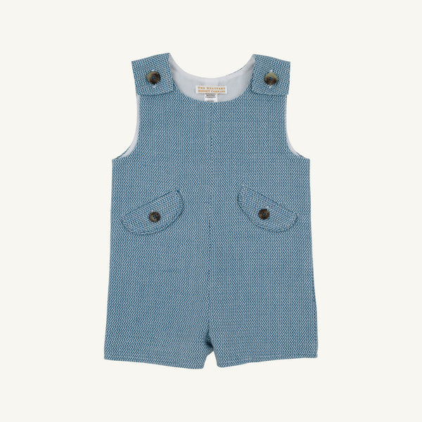 A sleeveless baby romper in a textured blue and white micro-pattern, featuring two front flap pockets with dark buttons and adjustable shoulder straps with metal snap buttons, shown flat on a light cream background.