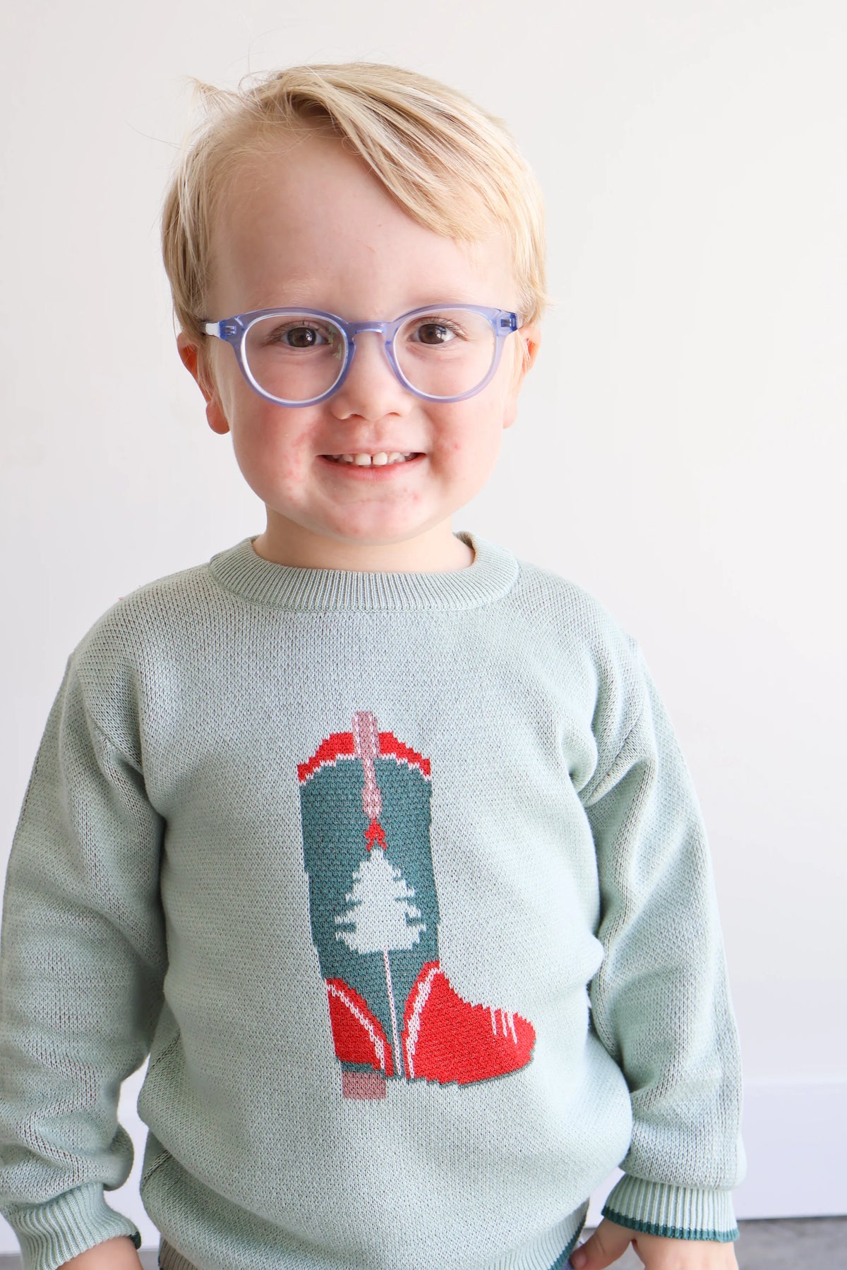 A young child with blonde hair and purple-framed glasses wearing a light sage green knit sweater with a red and teal cowboy boot design on the front, set against a plain white background.
