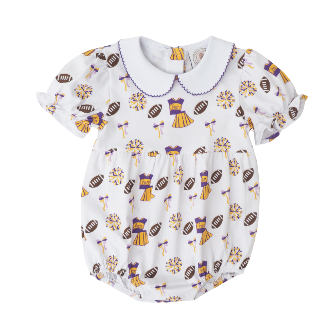 A baby short-sleeve bubble in white fabric with an all-over print of purple and gold football-themed illustrations, including footballs, cheerleaders, and bows. It features a white Peter Pan collar with purple trim and ruffled sleeves.