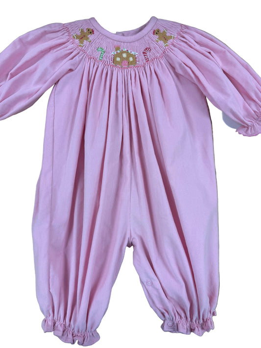 A baby pink long-sleeve footless smocked jumpsuit with elasticized cuffs, laid flat on a wooden floor. The chest features smocked gingerbread house design 
