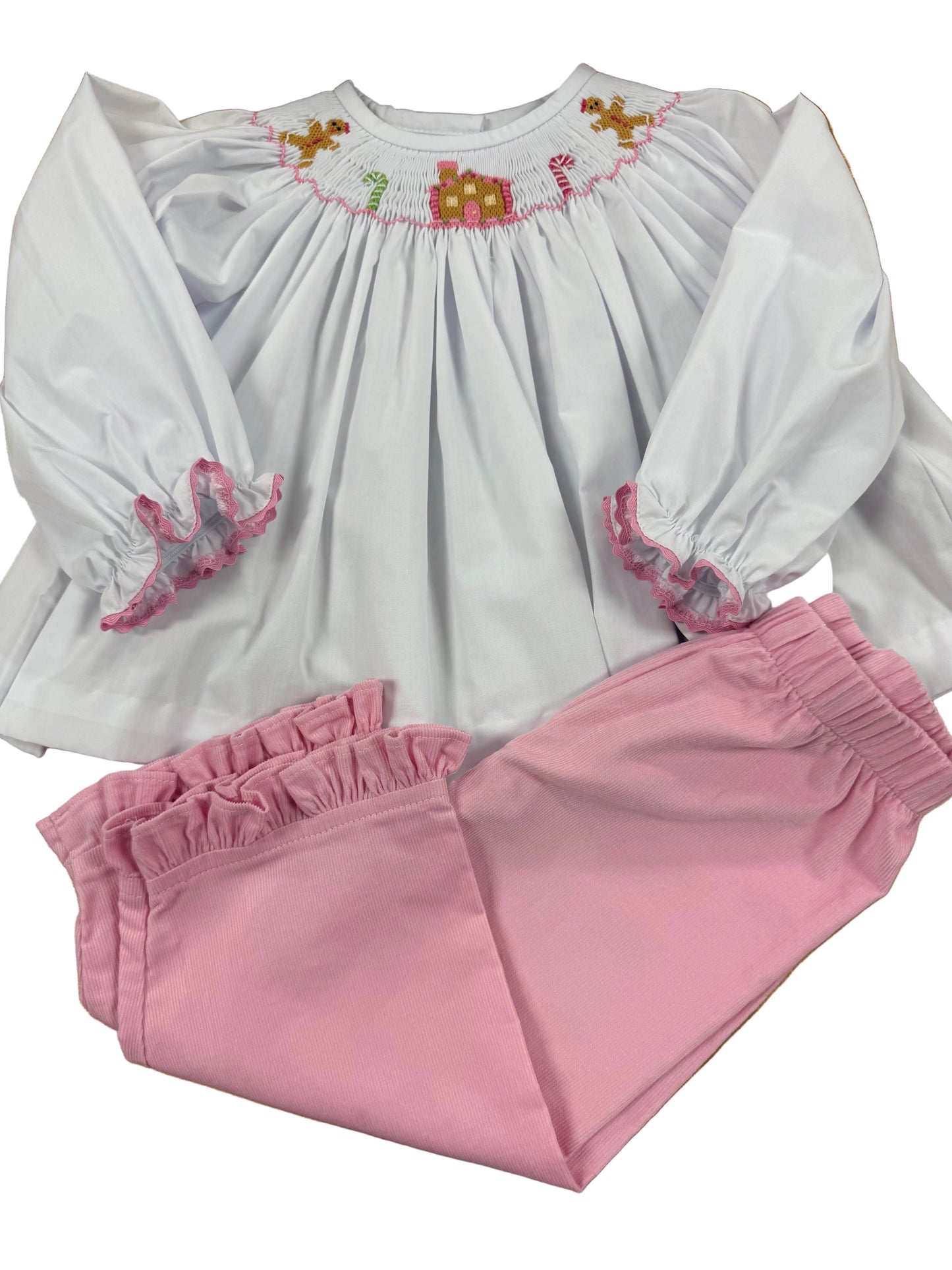 A children's two-piece outfit laid flat on a wooden floor, featuring a white long-sleeve smocked blouse with pink trim and gingerbread motifs at the neckline, paired with pink ruffled pants.