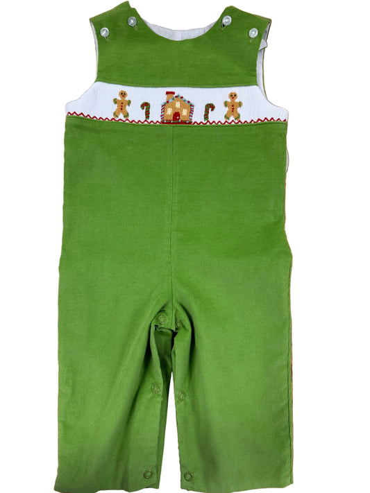 A green corduroy baby overalls romper with a white smocked chest panel featuring gingerbread men and a gingerbread house, laid flat on a wooden floor.
