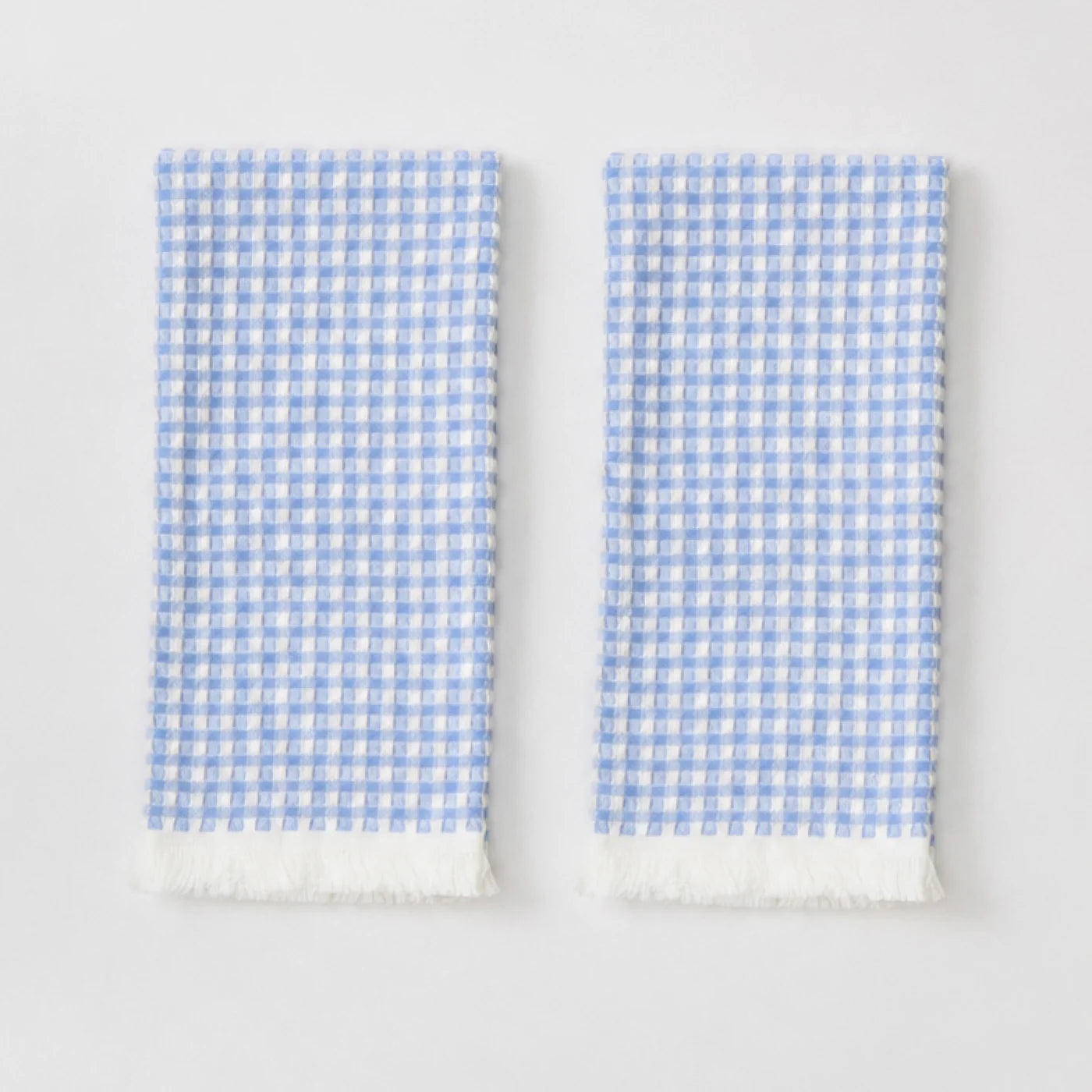 Two folded white kitchen towels with a light blue gingham check pattern and white fringed edges, displayed side-by-side on a clean white background.
