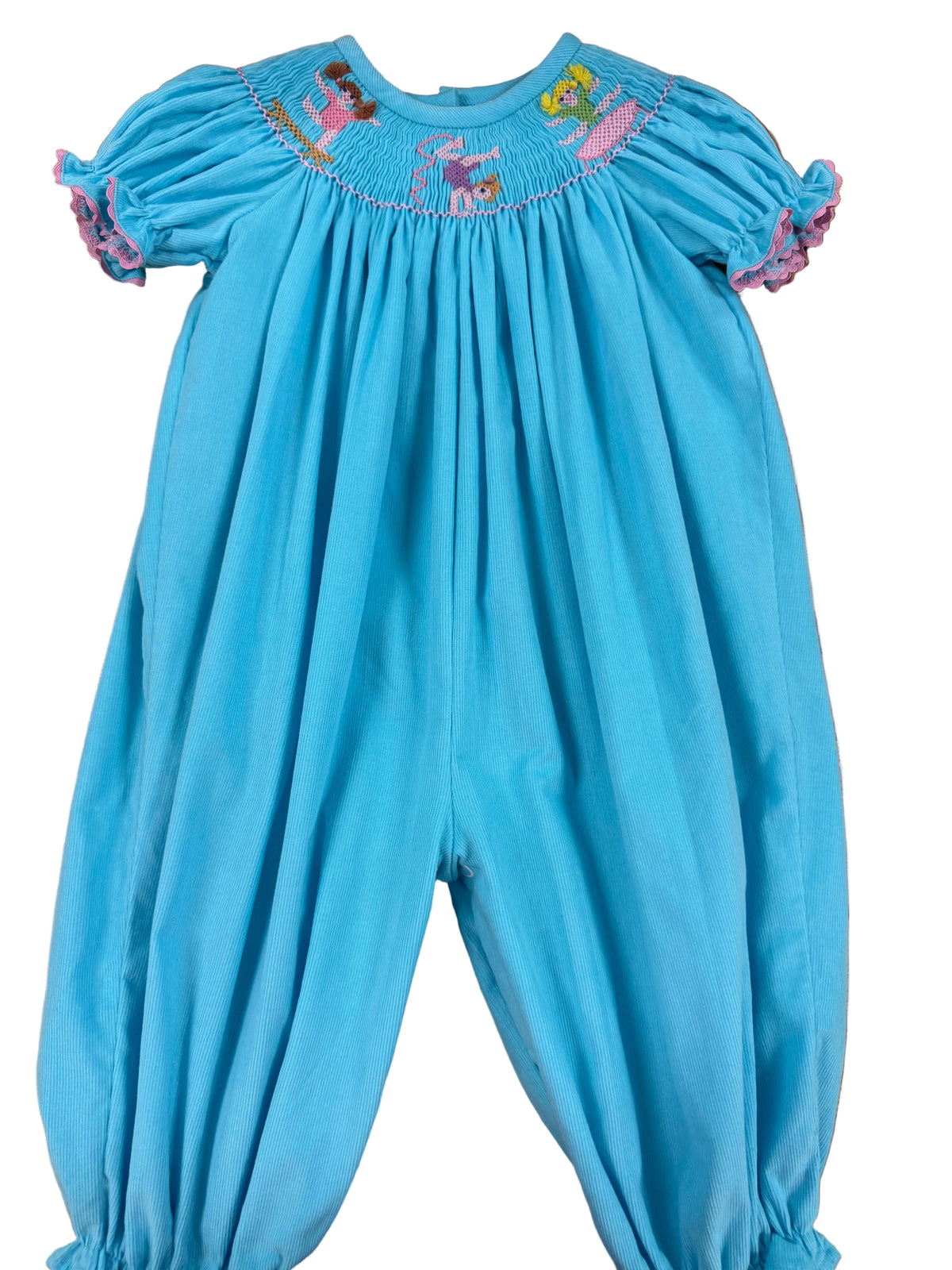 A baby smocked jumpsuit in bright turquoise blue, laid flat on a wooden floor. The jumpsuit features short puff sleeves with ruffled trim, a smocked yoke decorated with colorful embroidered figures, and elasticized leg cuffs.