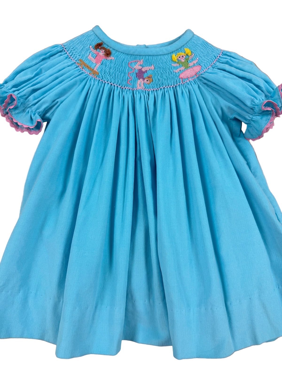 A light blue cotton smocked dress with short puff sleeves, decorated with embroidered gymnastics characters around the smocked yoke, laid flat on a wooden floor.