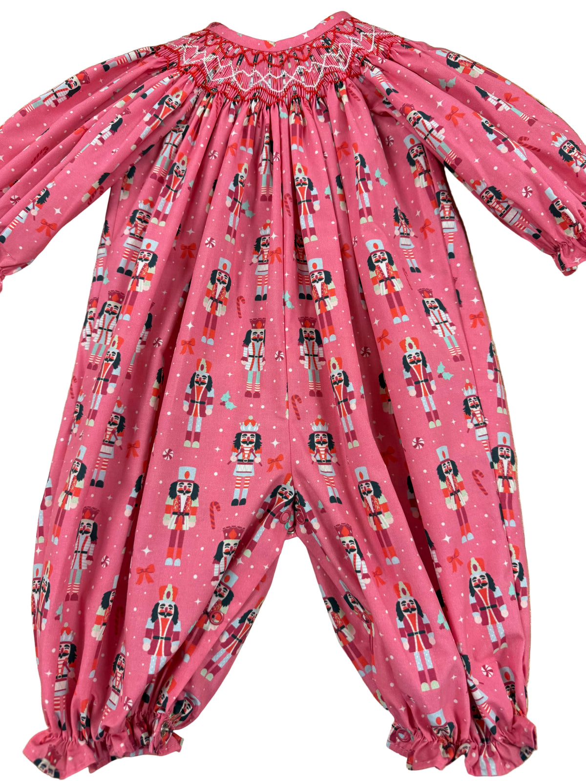 A pink long-sleeve baby jumpsuit with an all-over nutcracker print, laid flat on a wooden floor. The jumpsuit features a smocked neckline and elasticized ruffled cuffs on the legs.