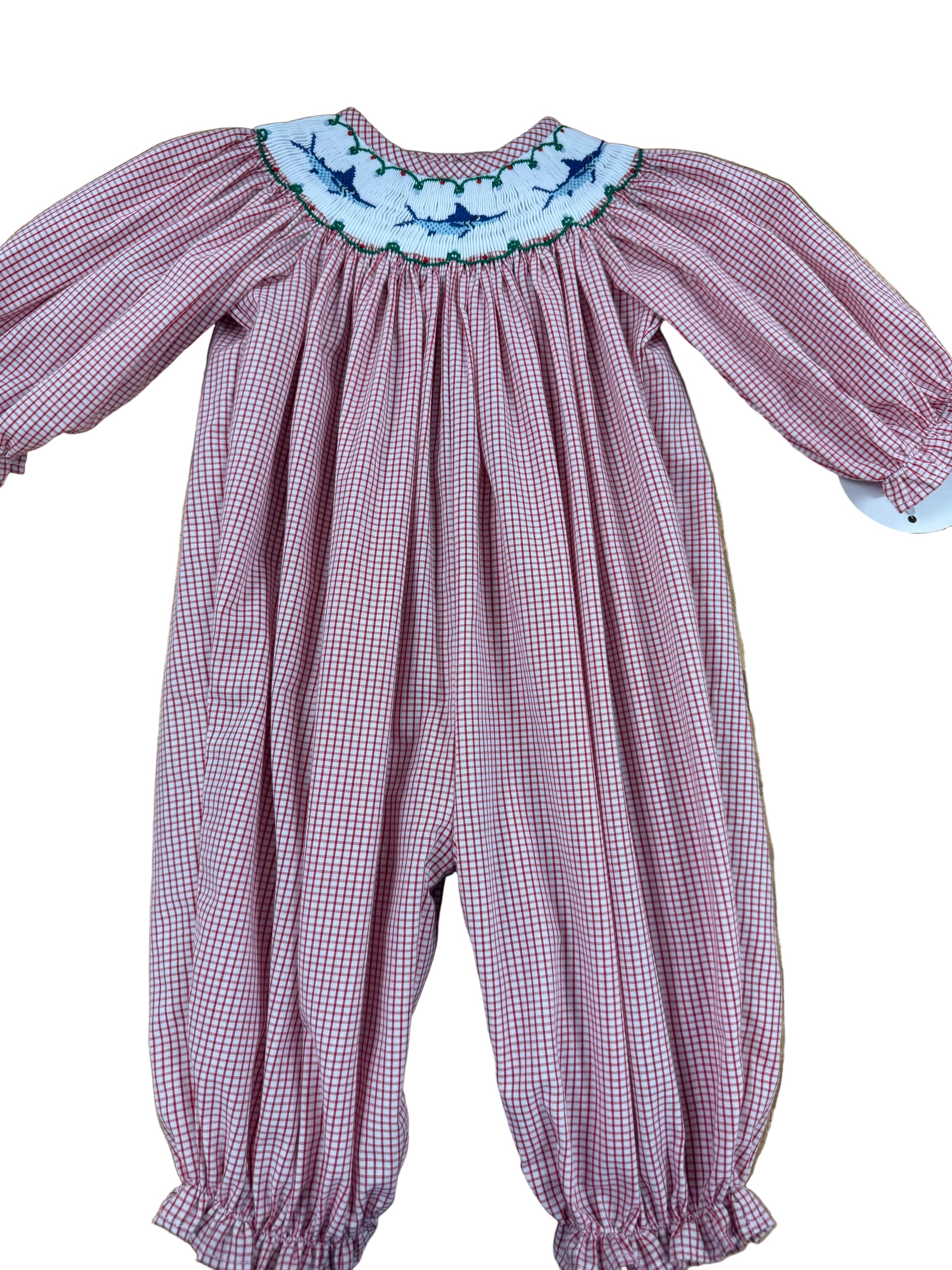 A baby smocked romper in a pink and white gingham pattern, featuring a white floral embroidered collar with blue and green accents, laid flat on a wooden floor.
