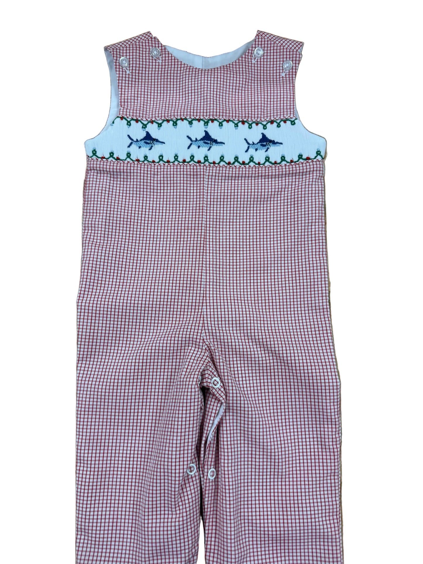 A baby's red and white gingham smocked overalls laid flat on a wooden floor. The chest features a white smocked band with embroidered blue fish and green leaves.
