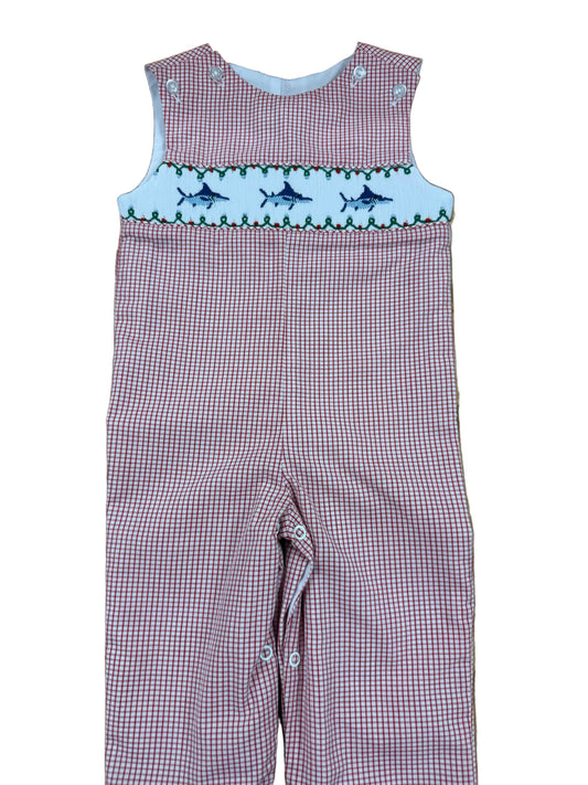 A baby's red and white gingham smocked overalls laid flat on a wooden floor. The chest features a white smocked band with embroidered blue fish and green leaves.