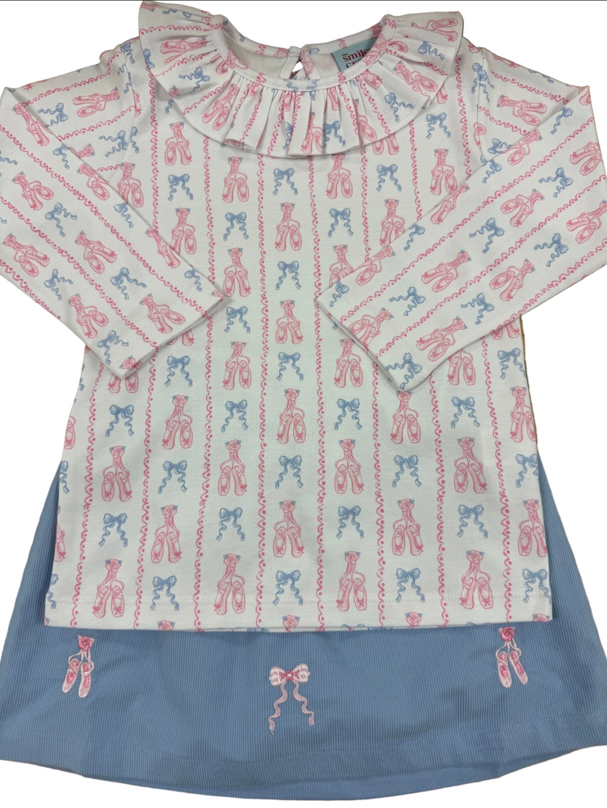 A children's long-sleeve top with a ruffled collar, featuring an all-over vertical pattern of pink and blue ballet pointe shoes on a light cream background, layered over a dusty blue skirt.