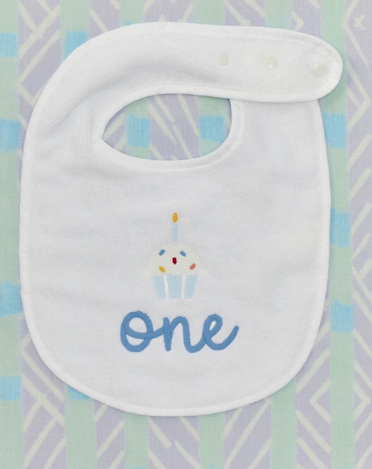 A white baby bib with a cupcake graphic and the word 'one' in blue script, laid flat on a pastel patterned background.