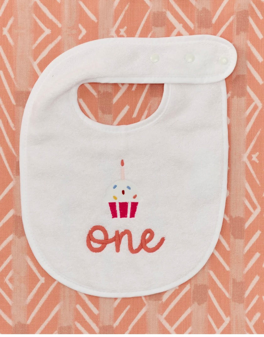 A white baby bib with an embroidered cupcake and the word 'one' in red script, displayed on a peach and white patterned fabric background.