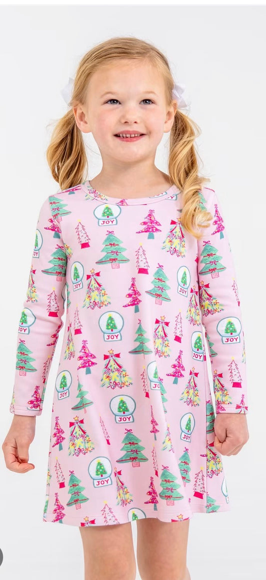 A young girl with blonde pigtails wearing a pink long-sleeve dress with a colorful Christmas tree pattern, standing against a plain white background.