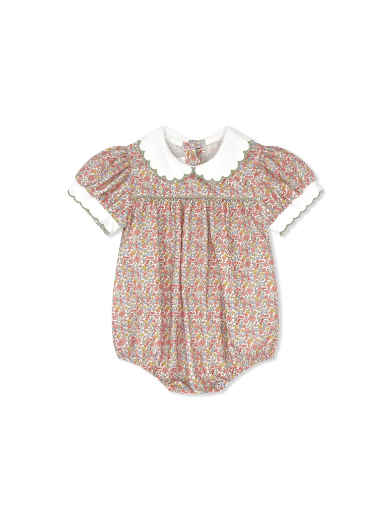 A baby romper with a dense multicolor floral print on a light pinkish-beige base, featuring a contrasting white Peter Pan collar and white ruffled short sleeves with green trim.