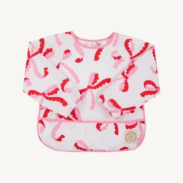 A long-sleeve baby bib with a white background and a pink and red abstract pattern, featuring a pink trim and a small circular gold logo patch on the lower right.