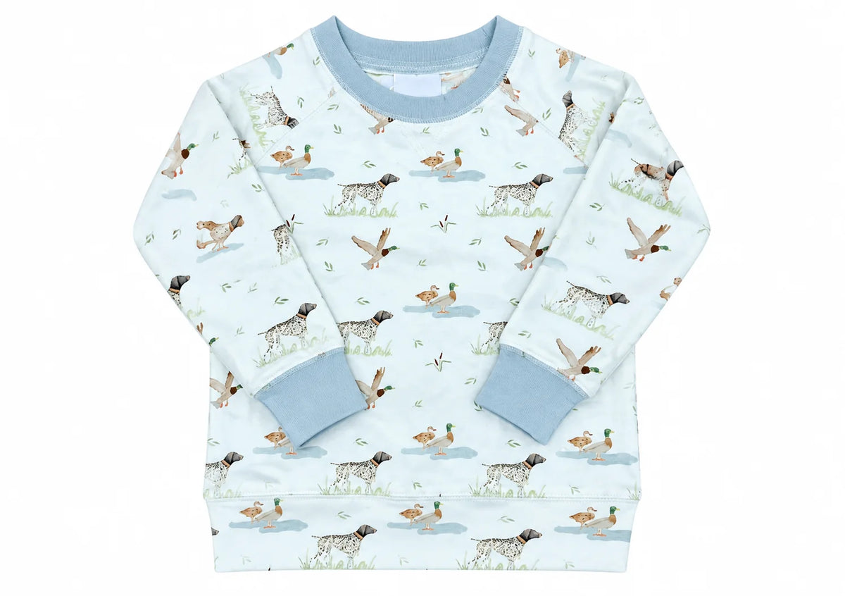 A long-sleeve baby sweatshirt featuring an all-over duck hunting print with dogs, ducks, and water elements on a cream background with solid light blue ribbed collar, cuffs, and hem.