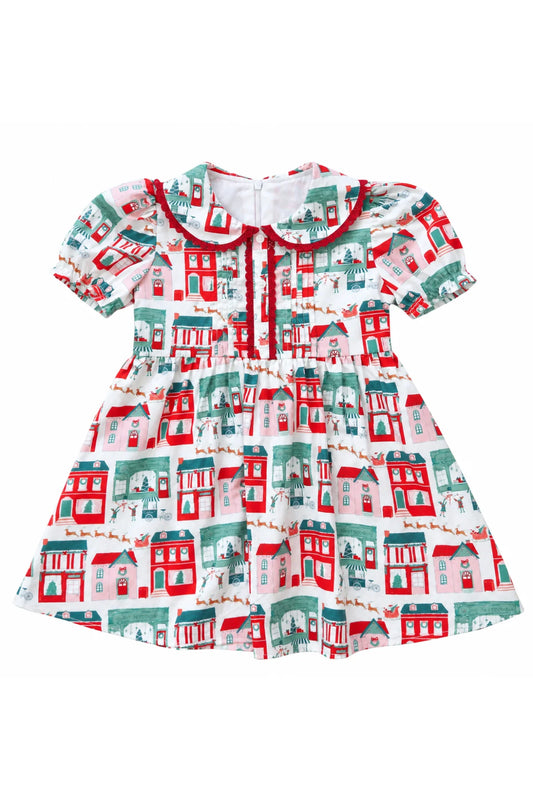 A short-sleeve baby dress with a white base and an all-over print of red and green houses, featuring a red collar and red tie detail at the neckline.