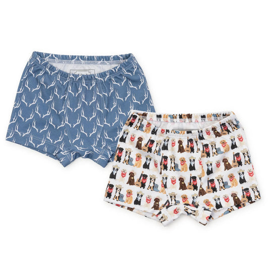 Two pairs of children's boxer shorts laid flat on a white background. The left pair is dusty blue with a white deer pattern, and the right pair is white with a repeating print of cartoon dogs in various colors.
