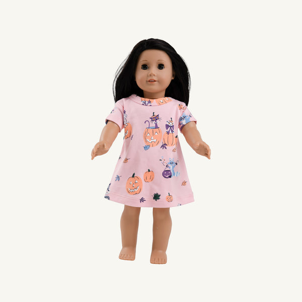A doll with long black hair wearing a light pink short-sleeve dress with a Halloween print of pumpkins, cats, and ghosts, set against a plain off-white background.