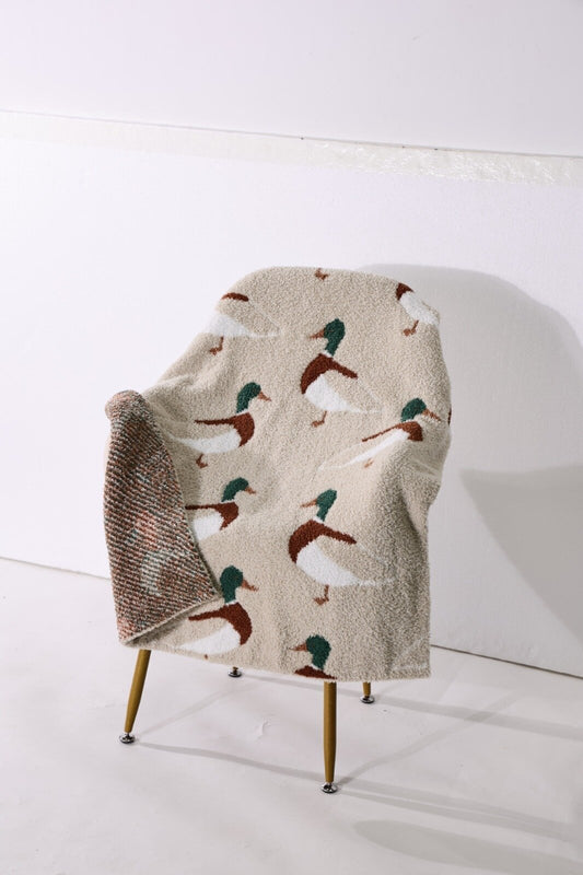 A beige fleece throw blanket with a repeating duck print in brown, white, and green, draped over a mid-century modern wooden chair against a white wall.