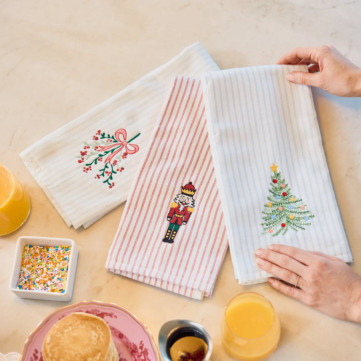 Three embroidered kitchen towels are displayed on a light beige countertop with breakfast props. The left towel is white with a pink bow and green holly design the middle towel is striped nutcracker and the right green striped with a christmas tree.