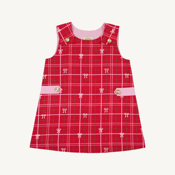 A sleeveless red plaid pinafore dress for toddlers, featuring a pink inner collar, pink button details, and a repeating pattern of small white bows on a light cream background.