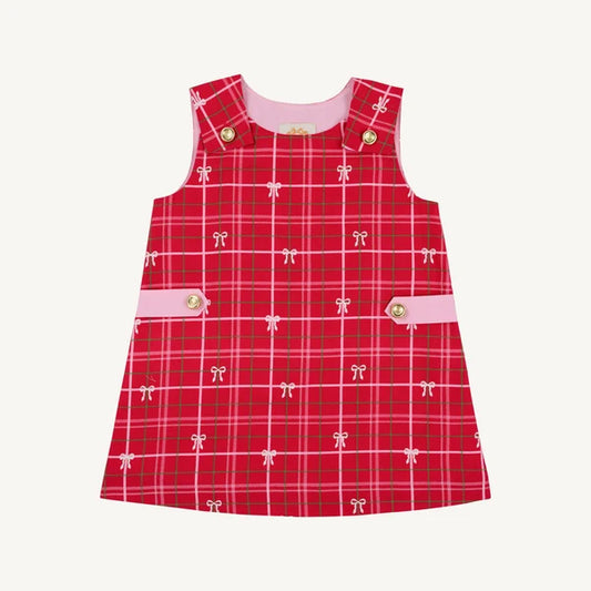 A sleeveless red plaid pinafore dress for toddlers, featuring a pink inner collar, pink button details, and a repeating pattern of small white bows on a light cream background.