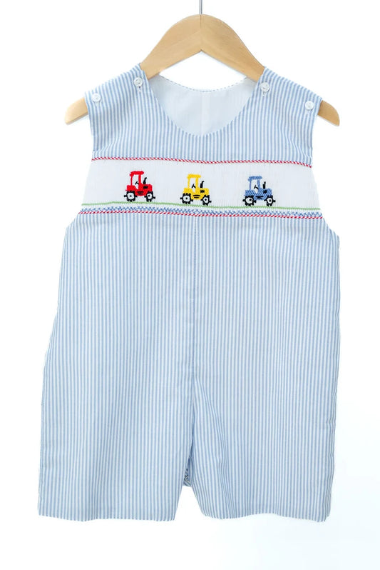 A baby boy's sleeveless smocked romper in light blue and white vertical stripes, featuring three embroidered toy tractors (red, yellow, and blue) across the chest. The romper is displayed on a wooden hanger against a clean white background.