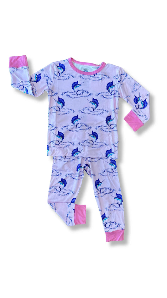A children's long-sleeve pajama set laid flat on a white background. The pajama top and pants are light pink with an allover print of blue and navy dolphins and waves, finished with bright pink cuffs and collar.