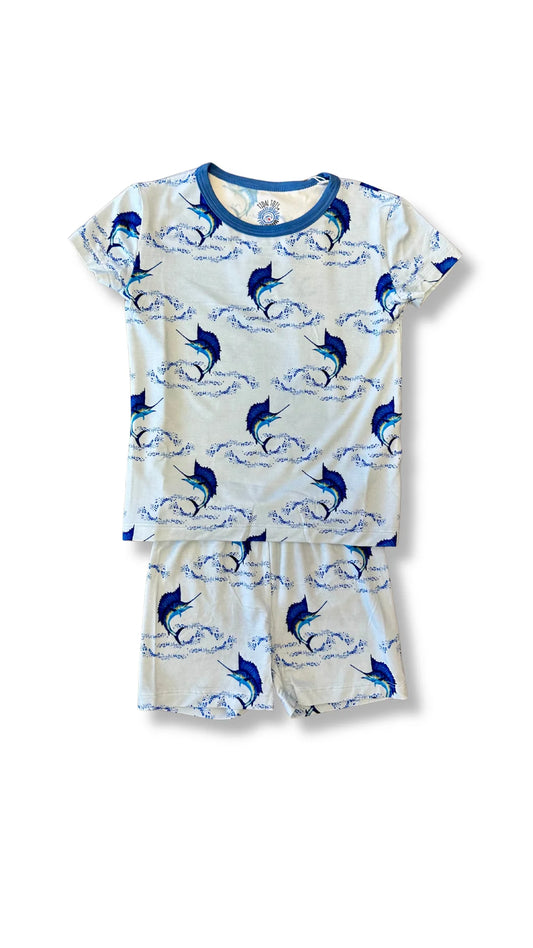 A two-piece children's pajama set featuring a short-sleeve top and matching shorts in an off-white color with a blue and navy print of jumping marlin fish and ocean waves. The top has a solid navy blue collar.
