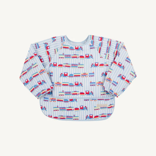 A long-sleeve baby bib with a light blue and white striped background and an all-over print of colorful toy trains. The bib has a wide underarm design and a small brand label on the lower right front.