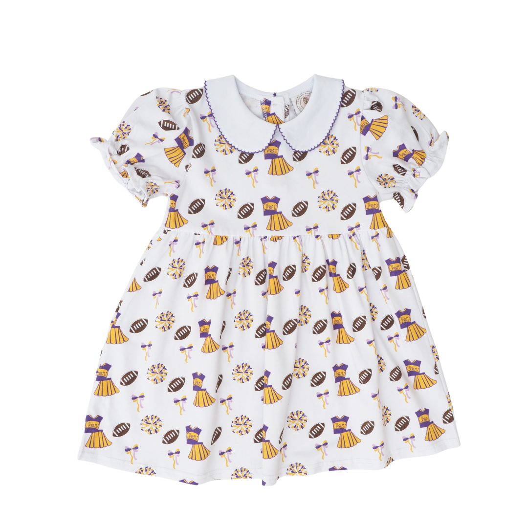 A white short-sleeve baby dress with a Peter Pan collar, featuring an all-over print of purple and gold football-themed illustrations including touchdowns, footballs.